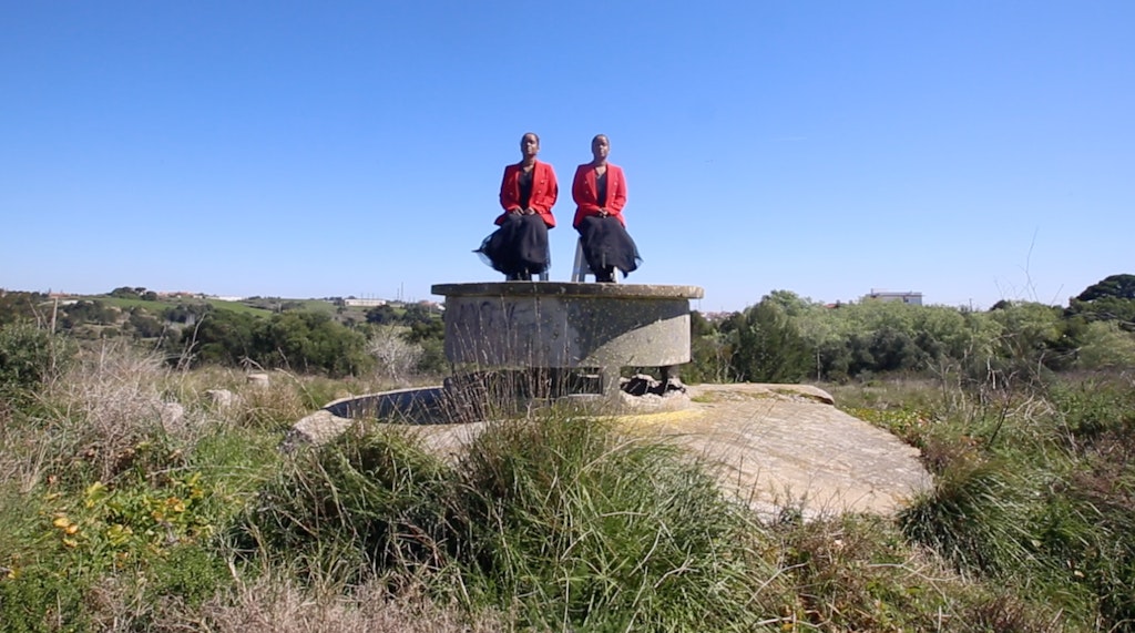Atop a concrete structure, two people in black dresses and red coats sit in chairs above the horizon of a grassy landscape.