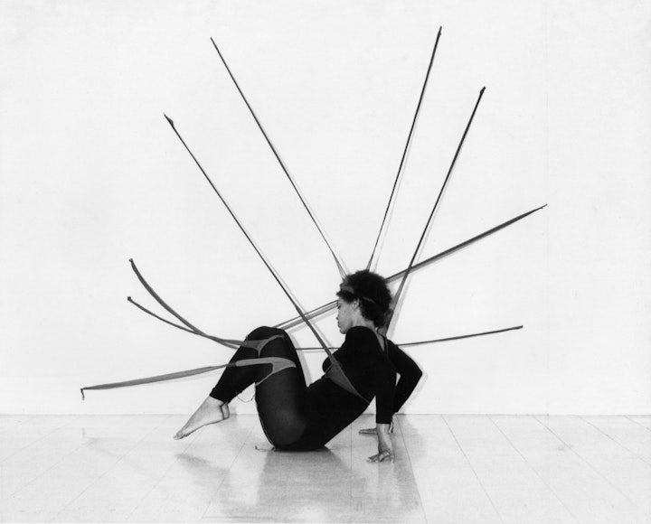 Black and white photo of a dancer performing in a gallery. They are wearing a black leotard and tights, sitting on the floor, wrapped in fabric strips which are attached to the gallery wall.