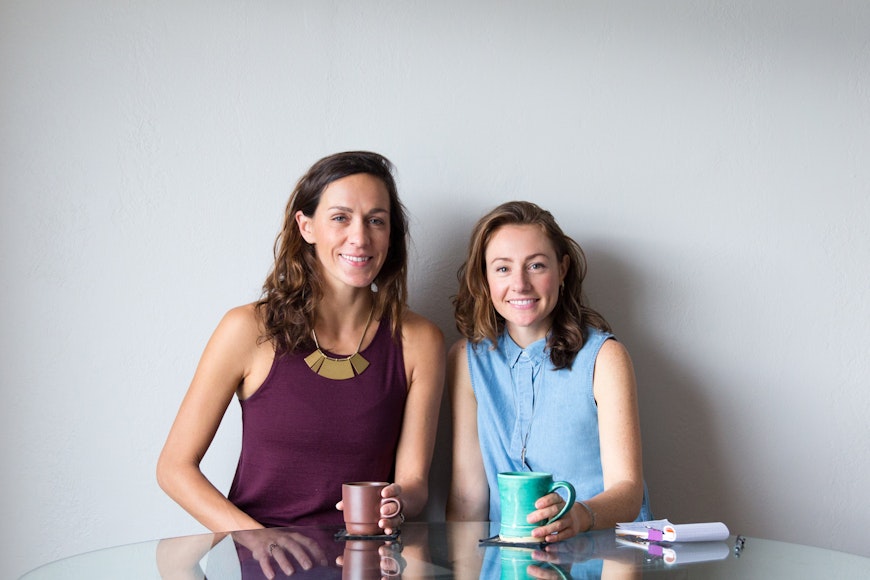 Portrait of two women against a white wall holding coffee mugs