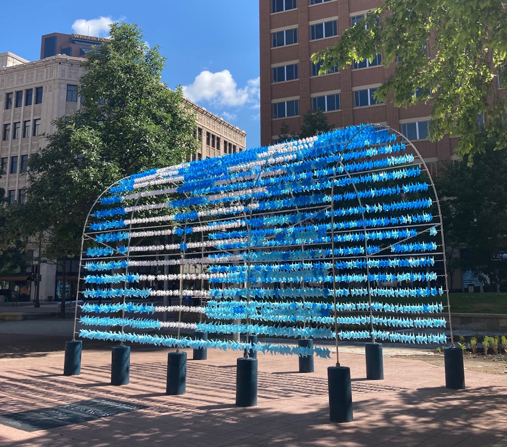 An archway in a courtyard with tall buildings in the background. The archway is tunnel-like, and made up of metal bars that are covered in small blue and white objects