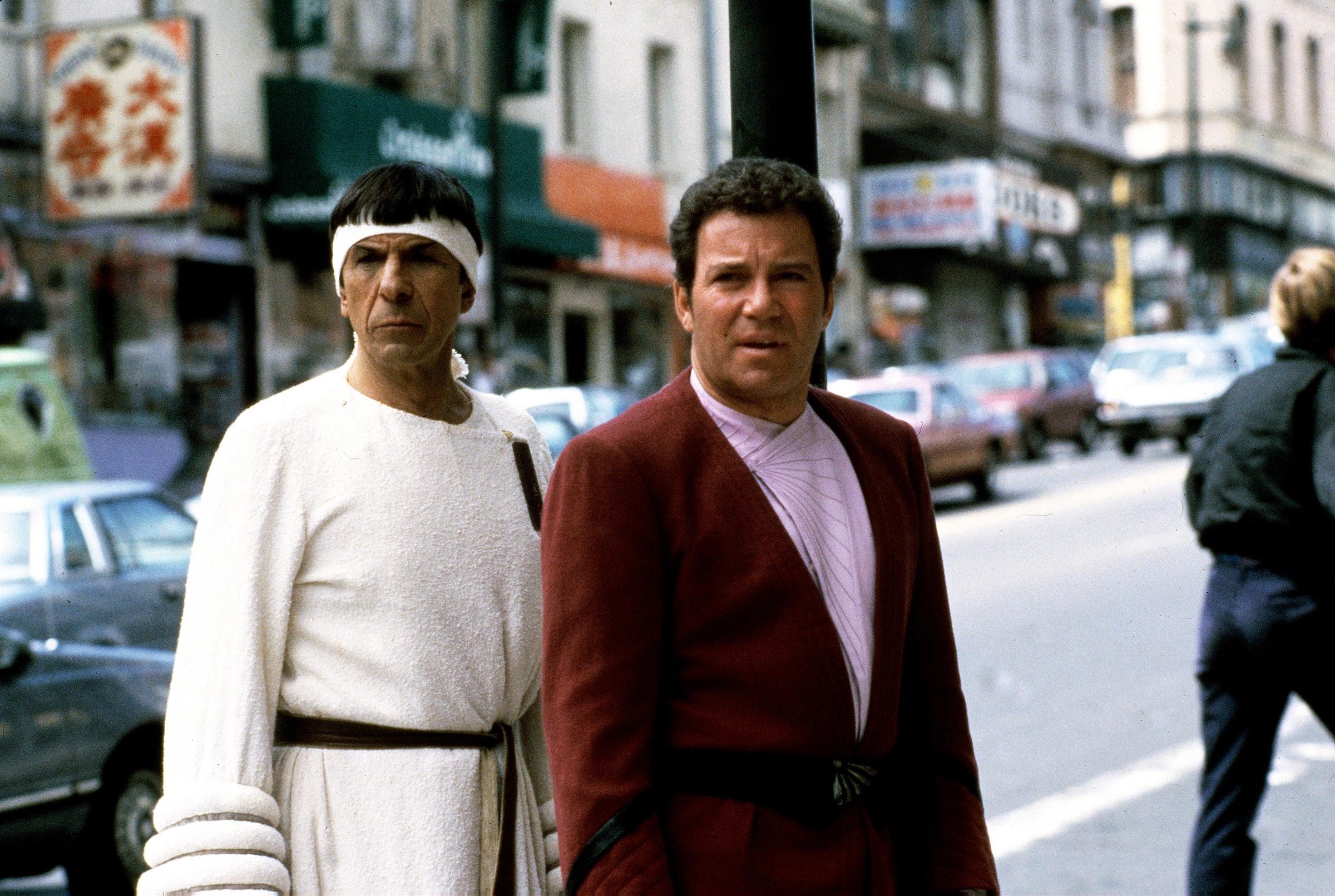 Two men on a chinatown street, Leonard Nimoy as Spock in a white robe and headband and William Shatner as Kirk in a red jacket and pink shirt.