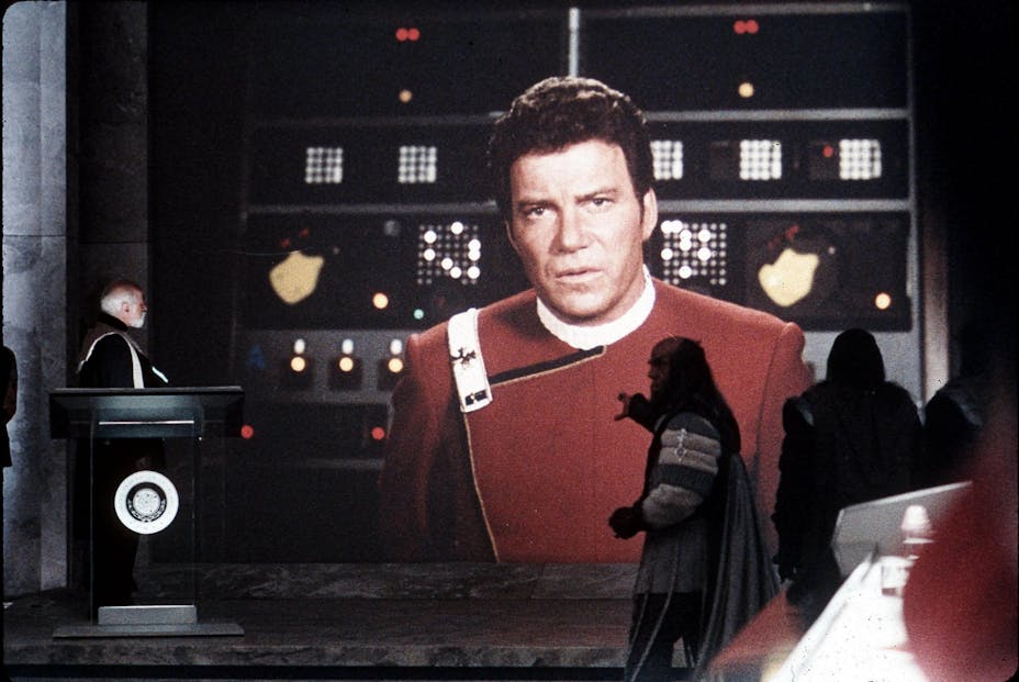 A man at a podium and a Klingon speaking and pointing, with a projection of Captain Kirk, a man with light skin and a red jacket, large behind them.