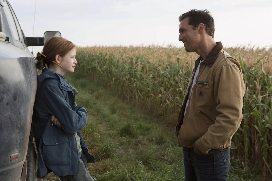 At the edge of a cornfield, a girl with light skin and red hair leans against a truck talking to her dad, standing and smiling, in jeans and a tan jacket.