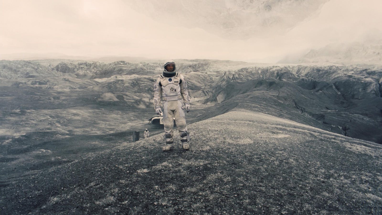 An astronaut stands on a hill in a snowy gray and black mountainous landscape with a white sky. Another astronaut is small in the distance.