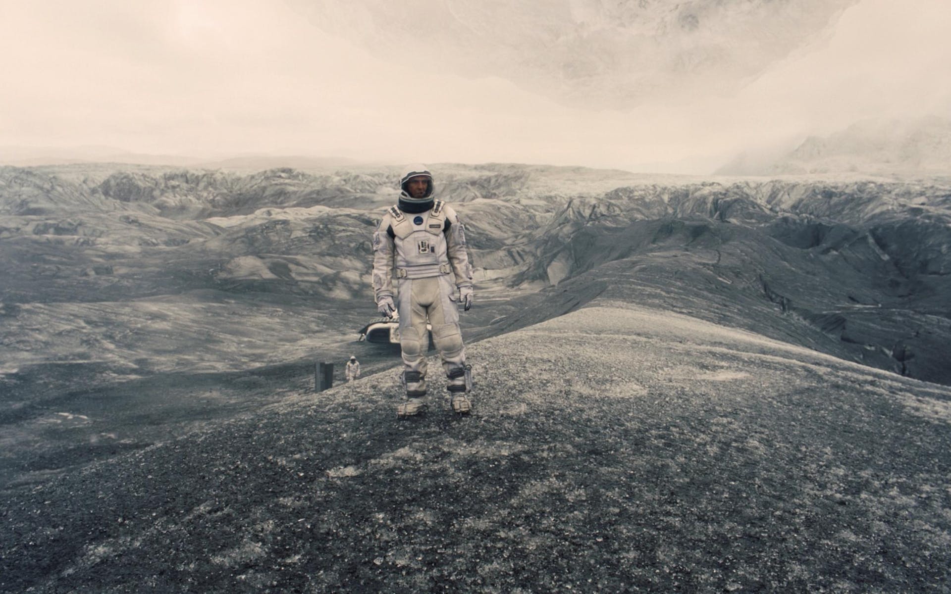 An astronaut stands on a hill in a snowy gray and black mountainous landscape with a white sky. Another astronaut is small in the distance.