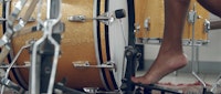 A close up shot of a medium-dark skin tone bare foot stepping on a kick drum pedal within a sparkly gold drum set.