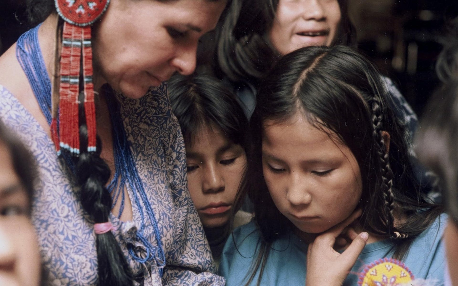 Alanis Obomsawin wearing a dress and beaded jewelry looks down at something in her lap with children around her looking on.