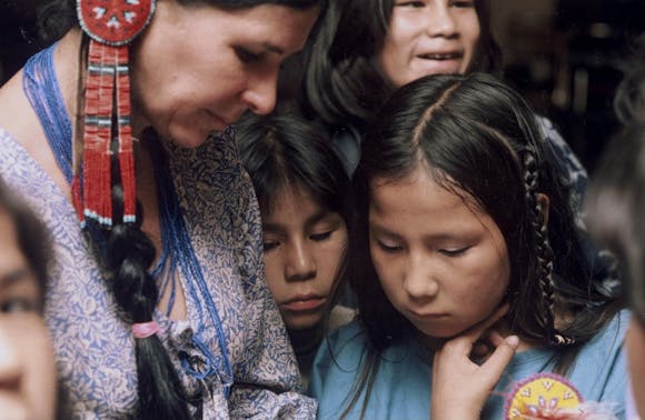 Alanis Obomsawin wearing a dress and beaded jewelry looks down at something in her lap with children around her looking on.