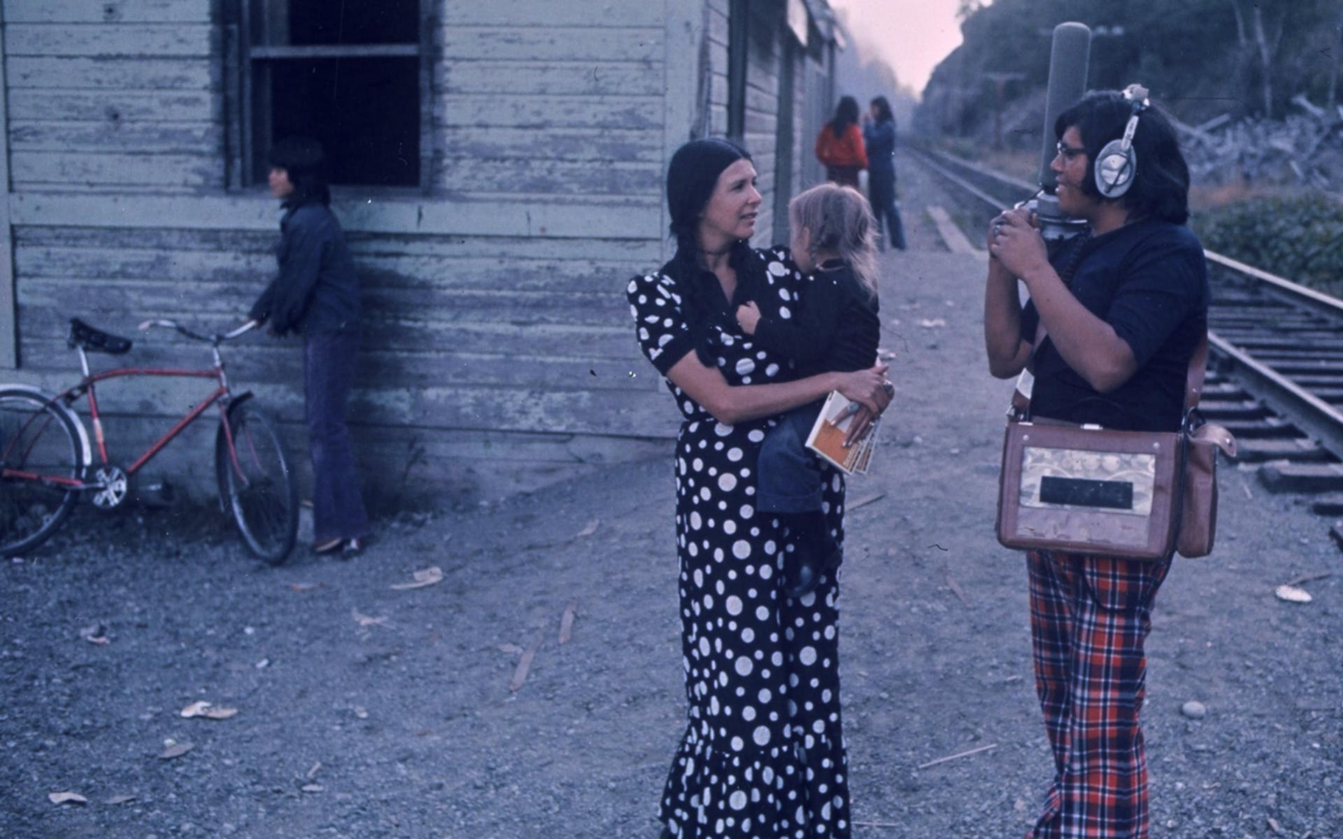 Alanis Obomsawin in a long polka dot dress holding a child and talking with a film crew member outdoors near train tracks.