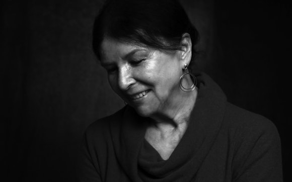 Black and white photo of Alanis Obomsawin smiling and looking down. Her dark hair is pulled back and she wears a sweater.