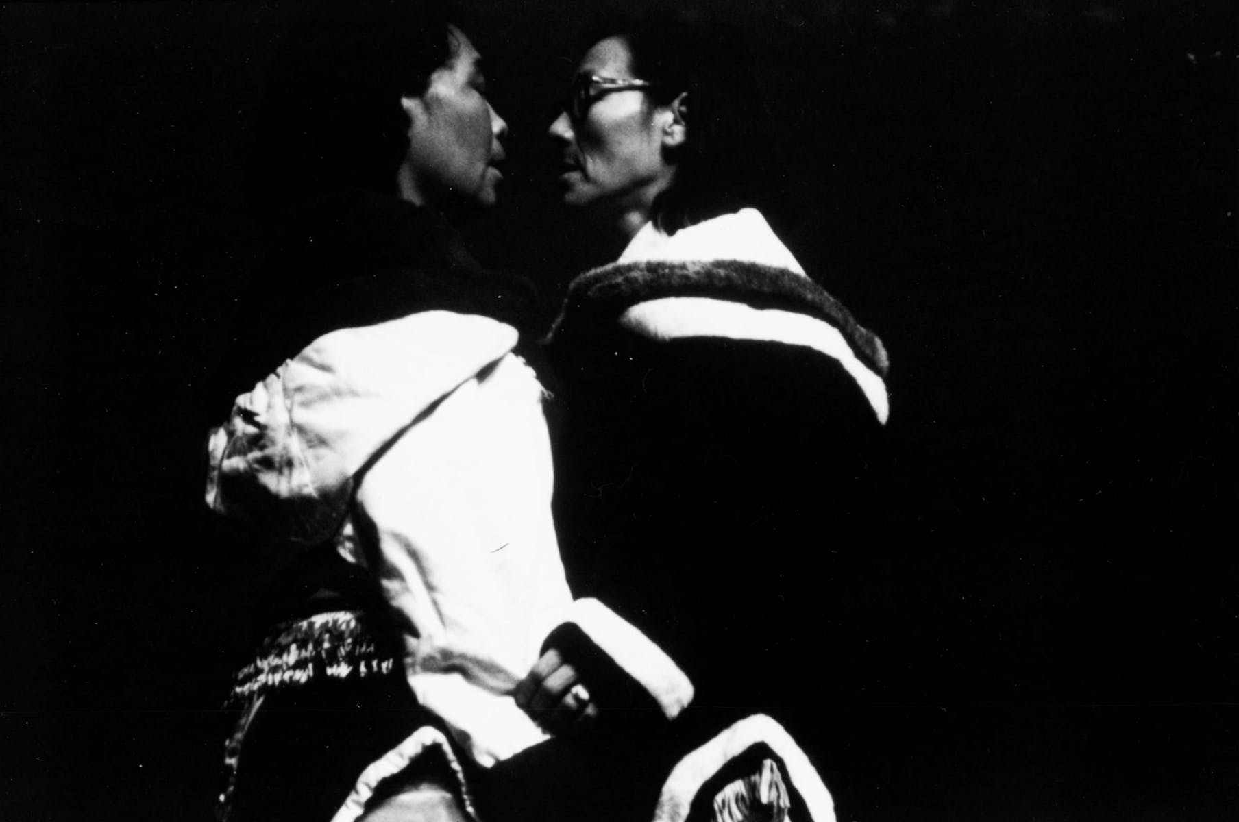 Black and white image of two people facing each other with their faces close together; it is high contrast and mostly black.