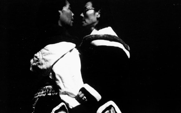 Black and white image of two people facing each other with their faces close together; it is high contrast and mostly black.