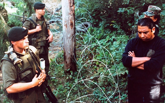 Outdoors amongst foliage, military personnel stand to the left of a razorwire barrier and indigenous men stand to the right.