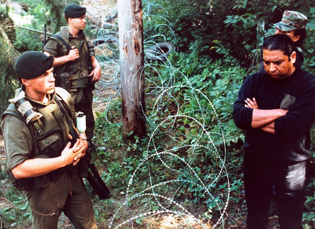 Outdoors amongst foliage, military personnel stand to the left of a razorwire barrier and indigenous men stand to the right.