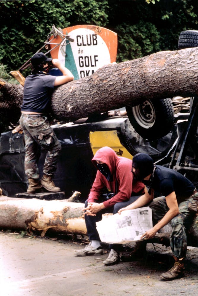 Three men with masks sit and stand on a barricade of massive logs and overturned vehicles beyond which a golf club sign is visible.