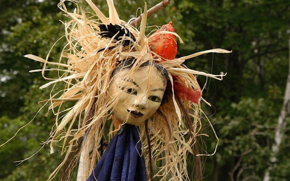 A handmade mask with the face of a woman surrounded with straw raffia, feathers, a gourd and purple fabric photographed outside.