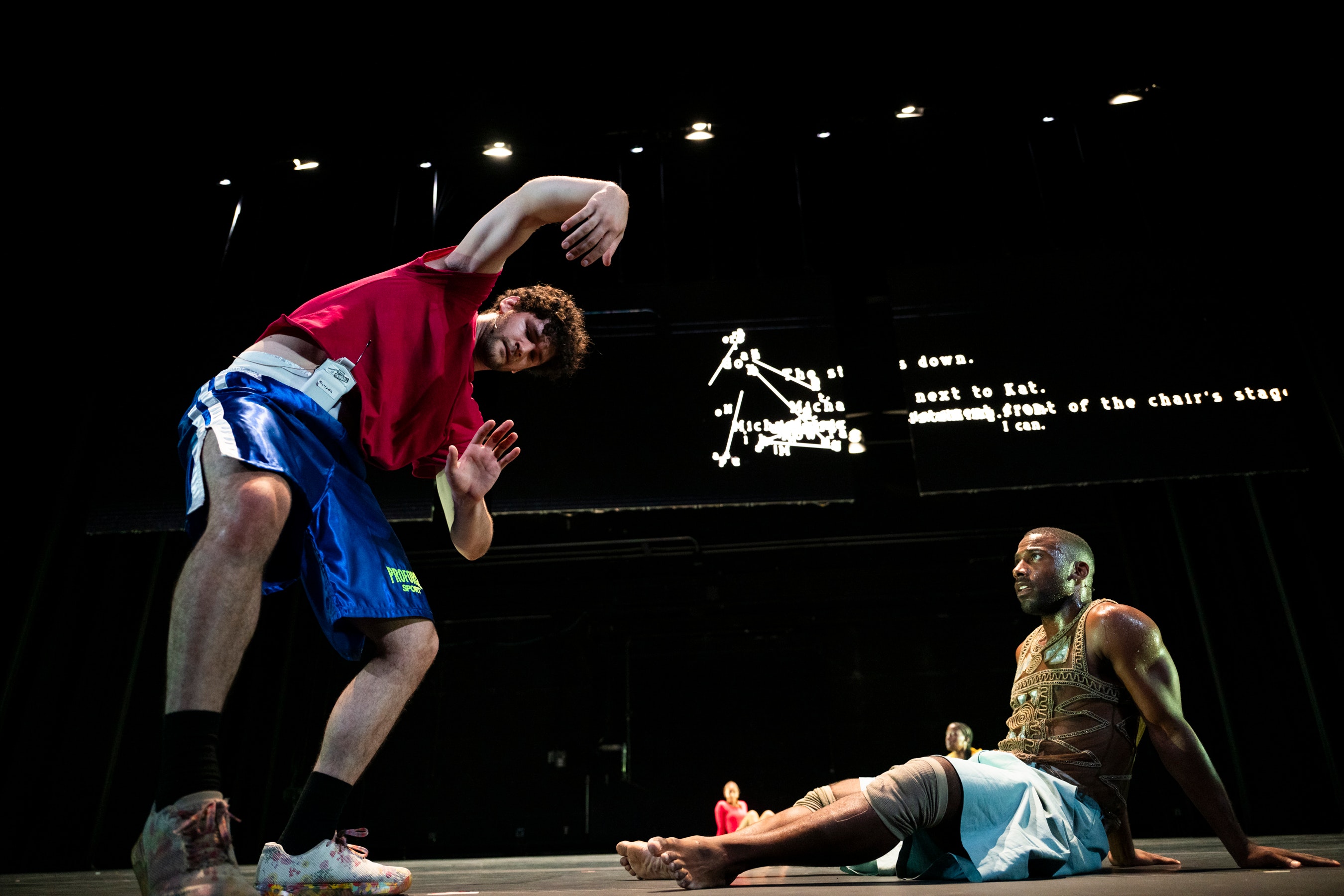 Two dancers—one standing, one sitting—shown in movement. Behind them is a screen with hard-to-read text.