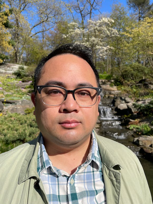 A man wearing glasses looks at the viewer while standing outdoors next to a stream and woods.