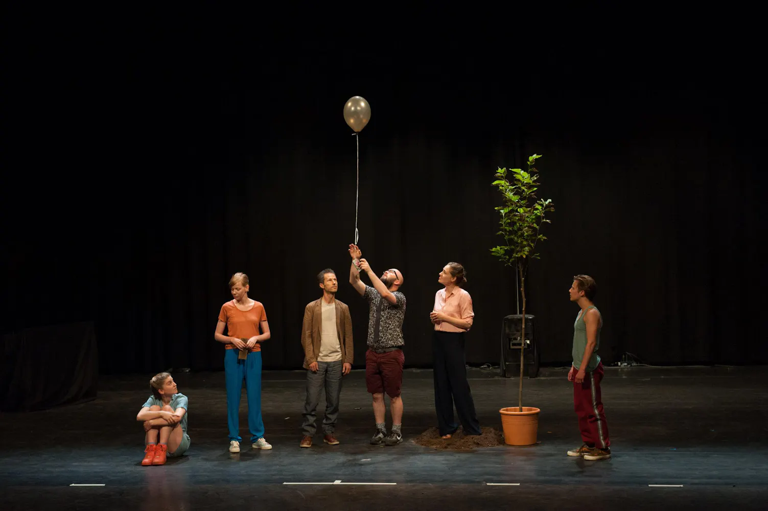 A group of adults stand on a dark stage with a potted tree while one of them holds a balloon on a string.