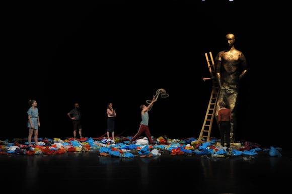 A group of adults stand ontop of many pieces of clothing on a dark stage while one of them climbs a ladder leaning against a large statue of a person.