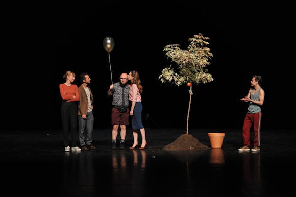 A group of adults stand on a dark stage with a potted tree while one of them holds a balloon on a string.