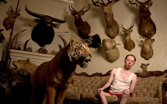 Man sitting on couch in front of a taxidermy tiger and several animal trophies hanging on the wall behind him.