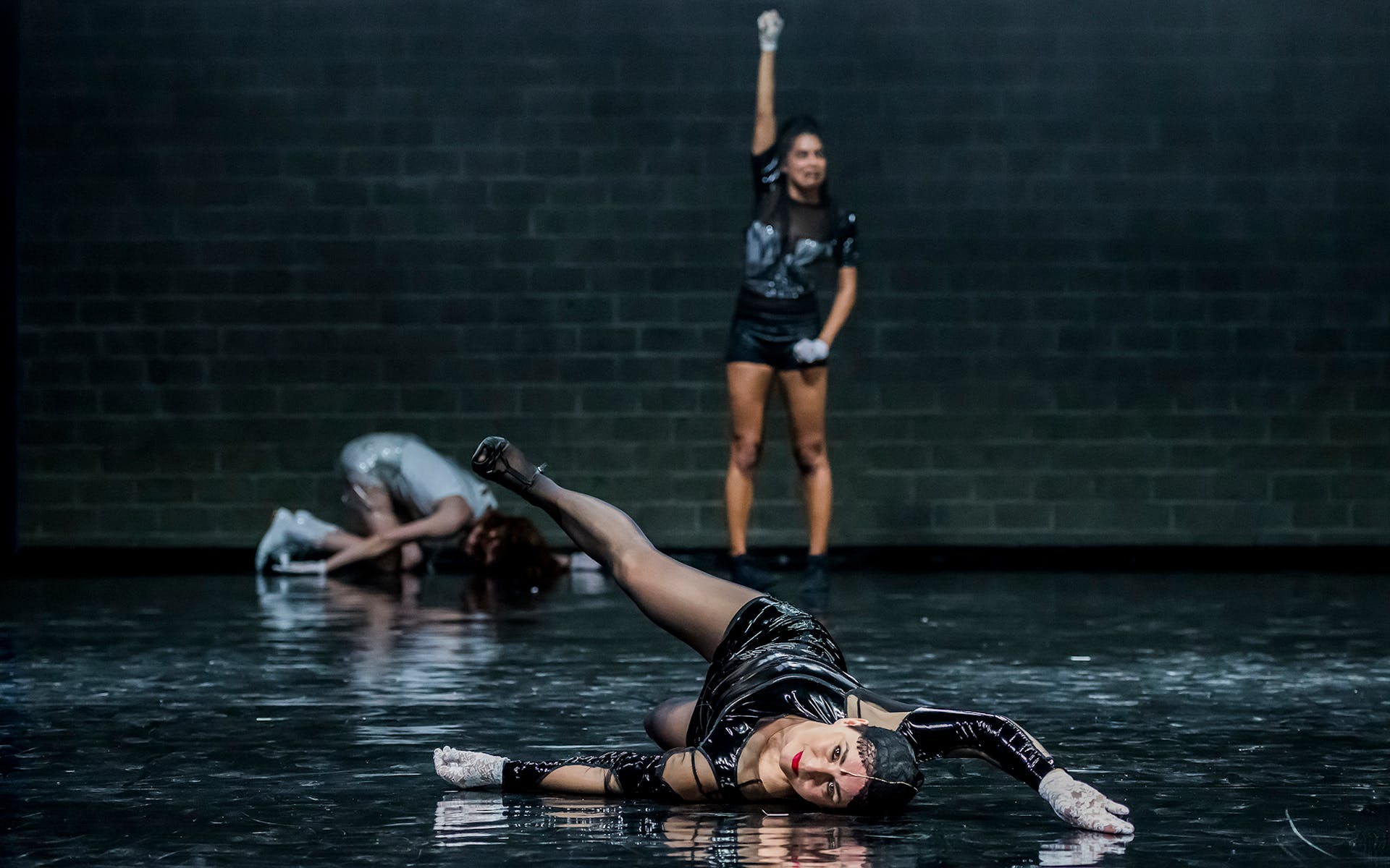 Two dancers on the ground, one standing with arm raised, on a black stage with brick wall background.