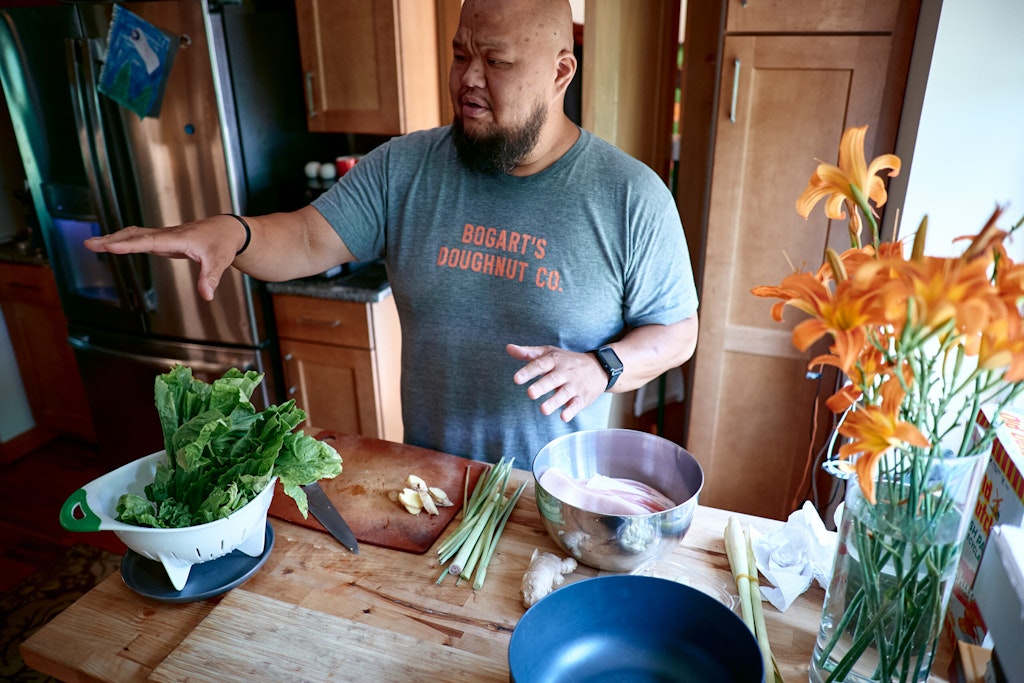 A man stands in a kitchen abd guestures at green onions, a bowl of greens, and other cooking items.