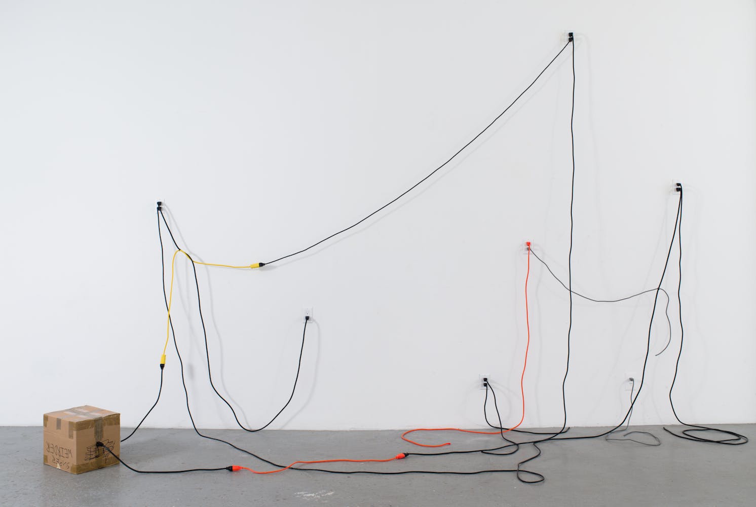 Various electrical cords of different colors plugged into outlets on the wall with a draping, sculptural effect