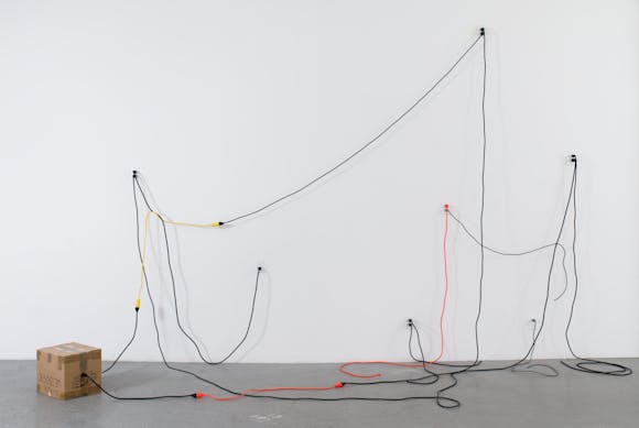 Various electrical cords of different colors plugged into outlets on the wall with a draping, sculptural effect