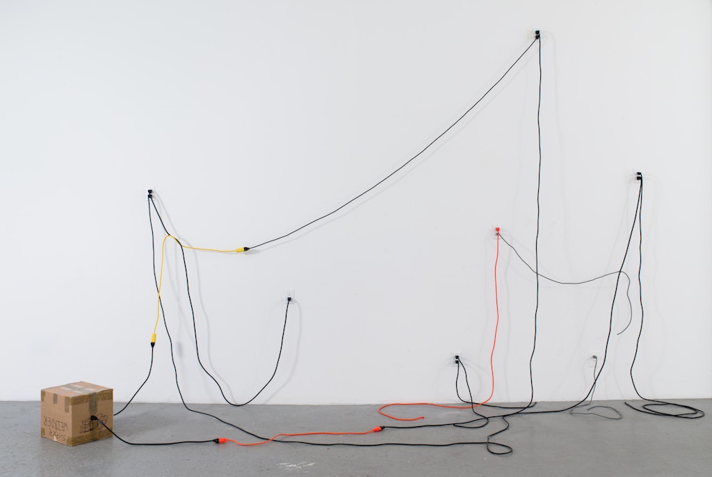 Various electrical cords of different colors plugged into outlets on the wall with a draping, sculptural effect