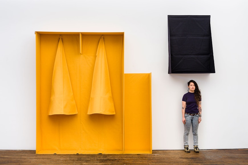An open box made of felt with two yellow cones hanging inside is next to a black felt box with a woman in a black t-shirt and jeans standing underneath.