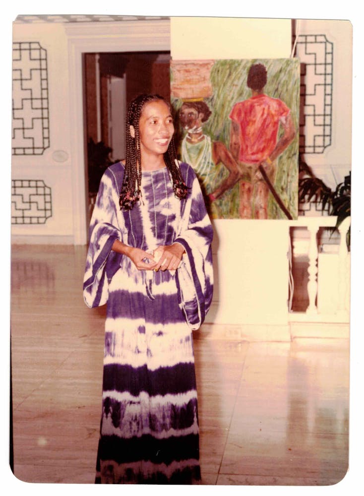 An adult woman smiles at someone offscreen as she stands in front of a painting of two figures in an art gallery.