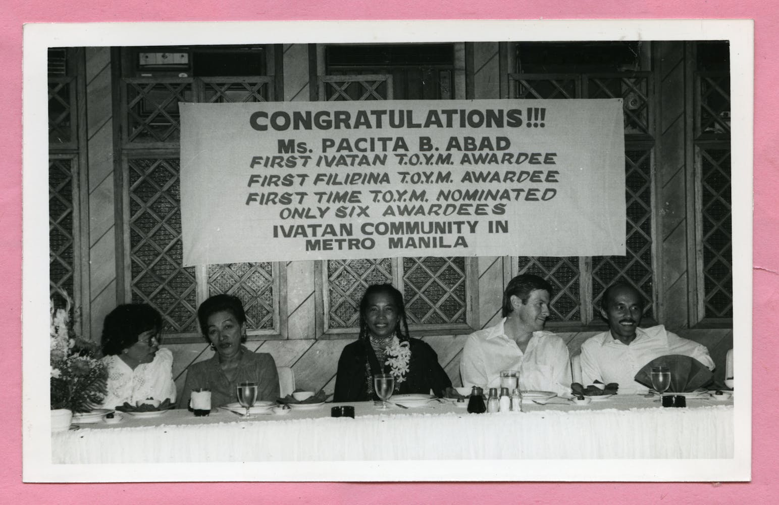 Several adults sit at a banquet table under a banner that congradulates Ms. Pacita B Abad.