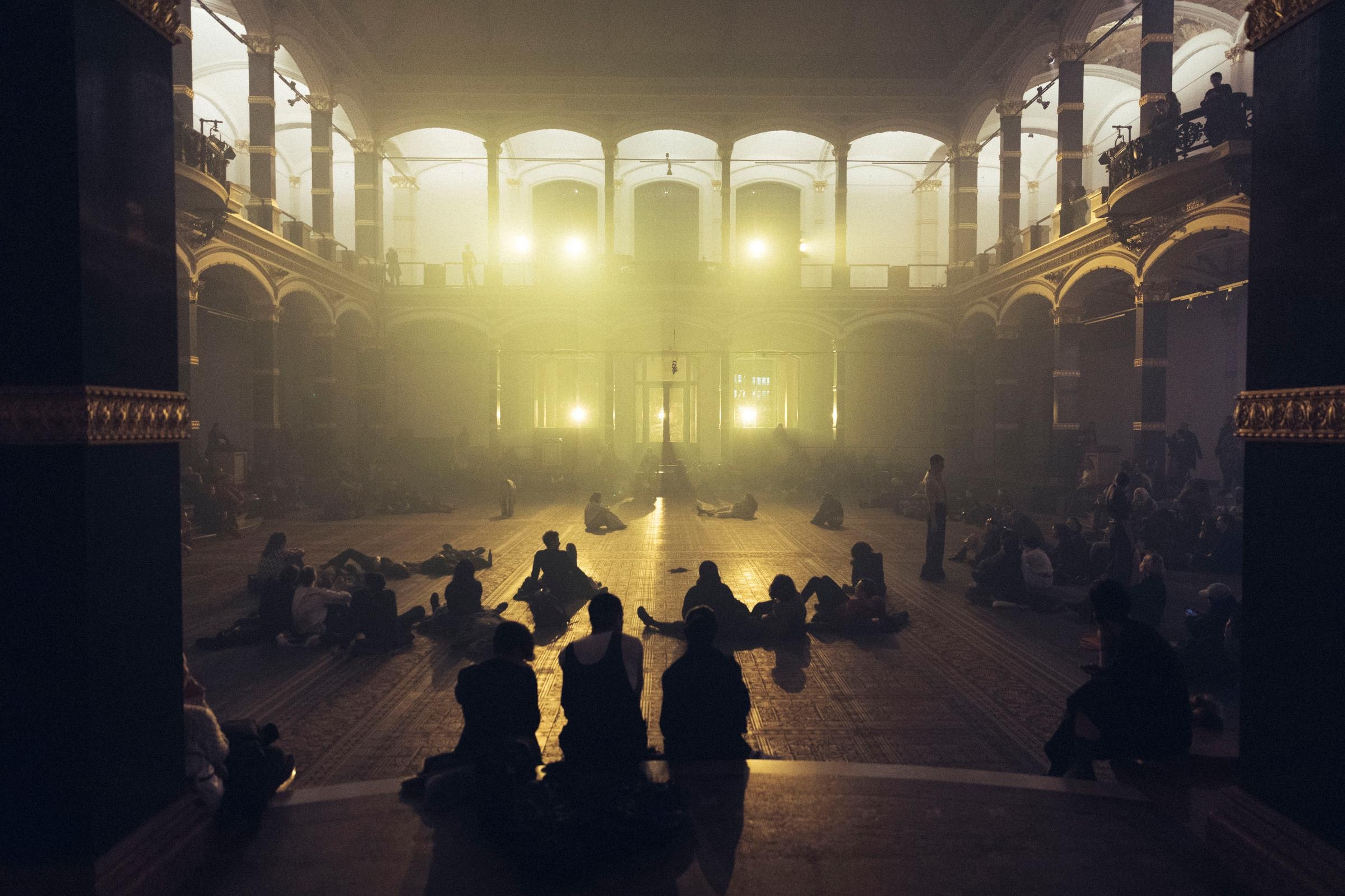 An audience and performers gathered in the lobby space of the Martin Gropius Bau in Berlin. Some are standing and others recline, all bathed in a yellow light.