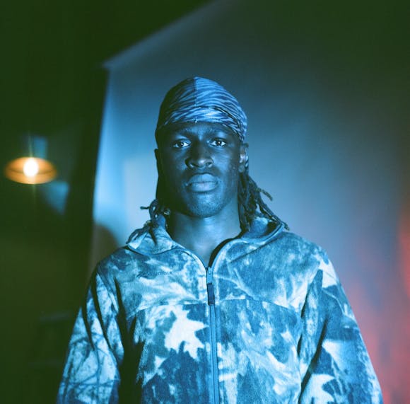 Photo of Papa Mbye, a man with medium-dark skin and shoulder-length dreadlocks, covered in blue, green, and purple light.