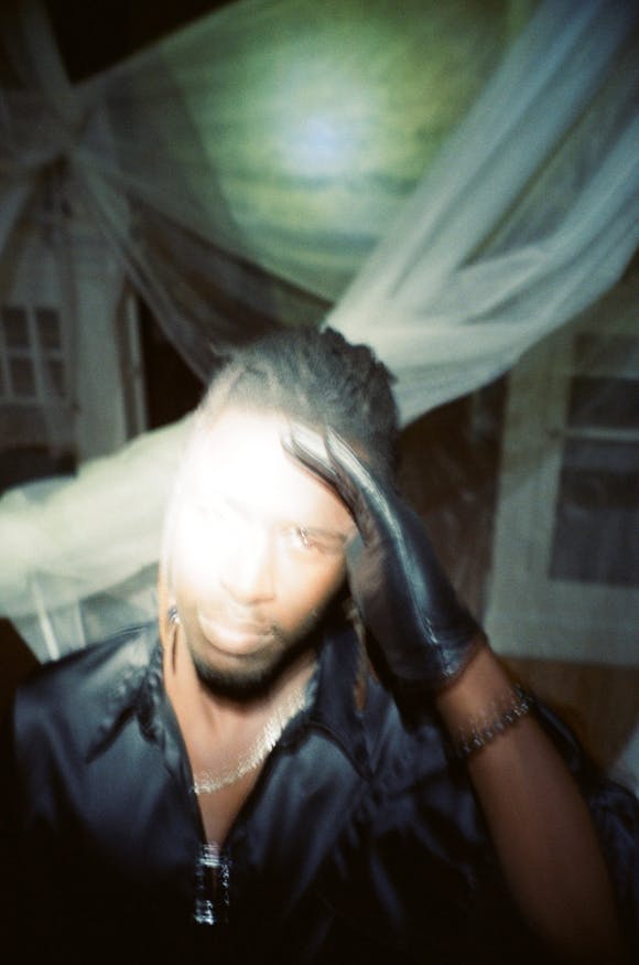 Blurred photo of Mbye wearing a black shirt and glove with his hand at his forehead looking into a bright camera flash.