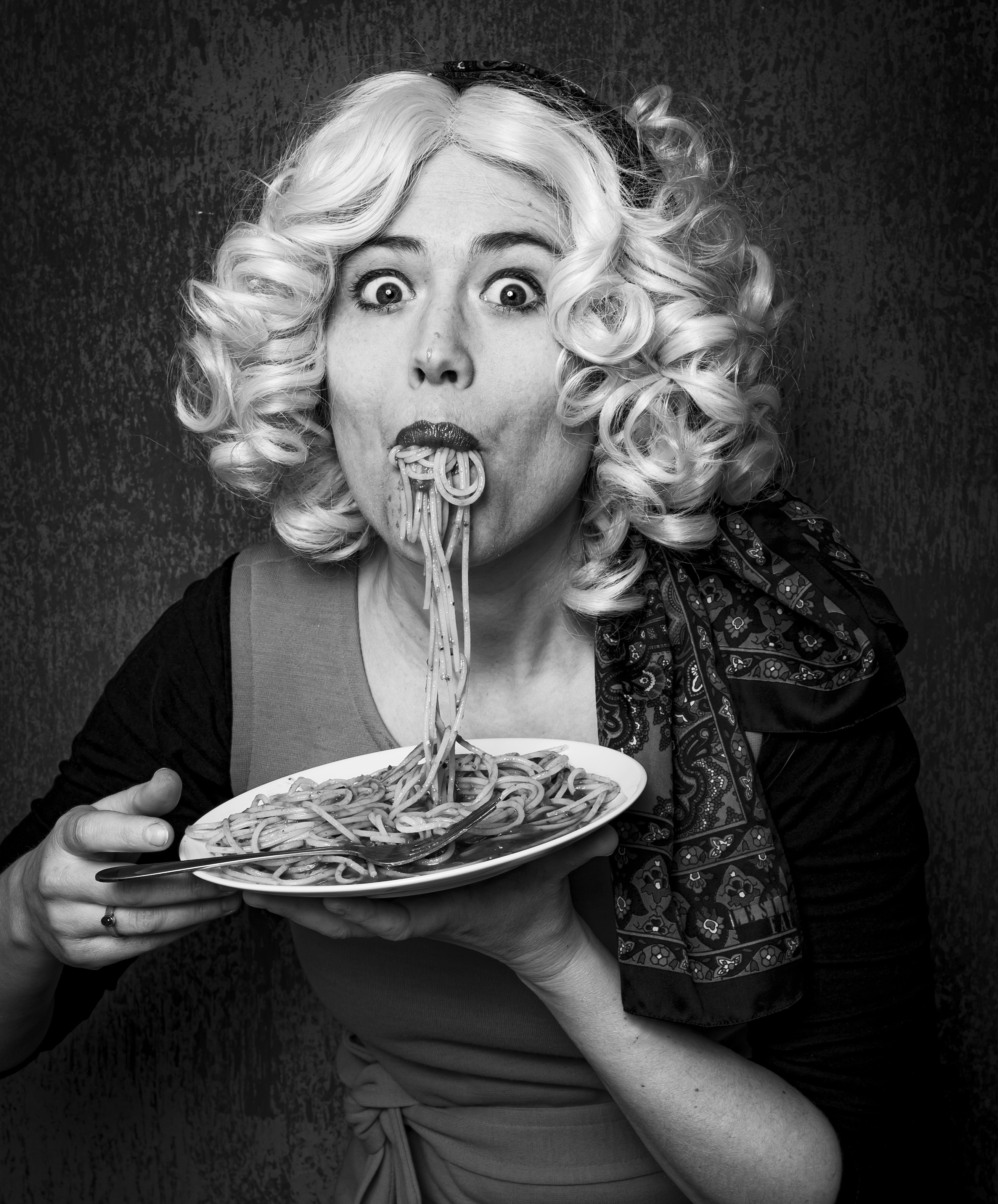 A woman dressed up with a mouthful of spaghetti overflowing out of her mouth onto a plate she is holding up. She has shoulder length curly hair and has her eyes open wide in a look of surprise