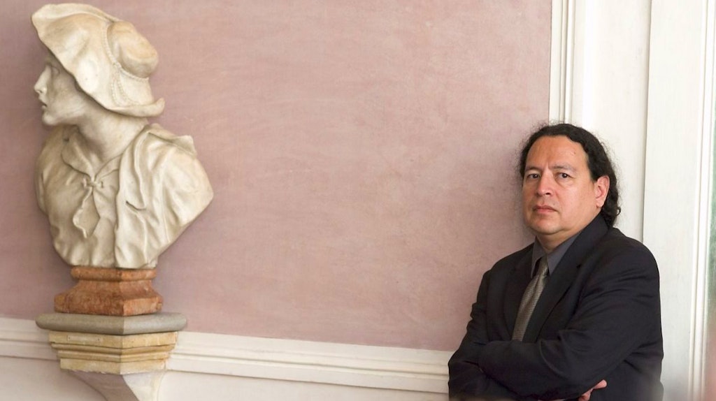 Paul Chaat Smith standing against a wall next to a bust of a woman.