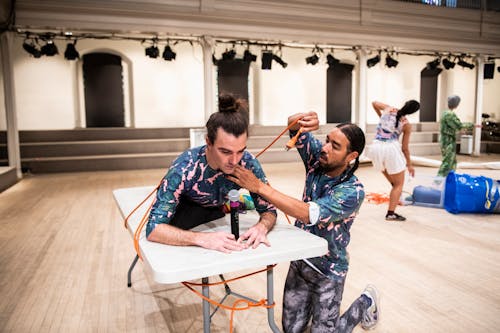 Silas leans forward while sitting on a collapsible table, like you would see at a picnic or a meeting room. Rashaun kneels on the floor beside the table, and has wrapped an orange extension cord around Silas.