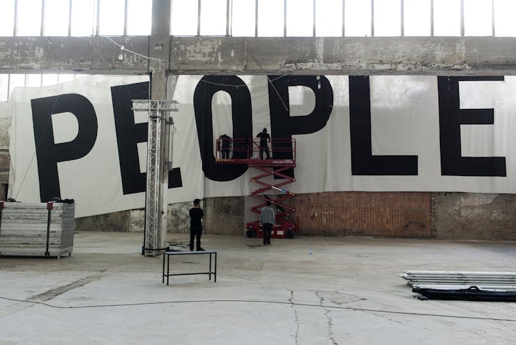 People hanging a large scale horizontal mural cloth mural that says "PEOPLE" in large black letters