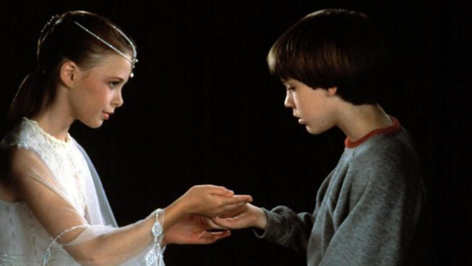 Two children with light skin and brown hair, a girl on the left in a white dress and a boy in a gray sweatshirt face each other, touching hands.