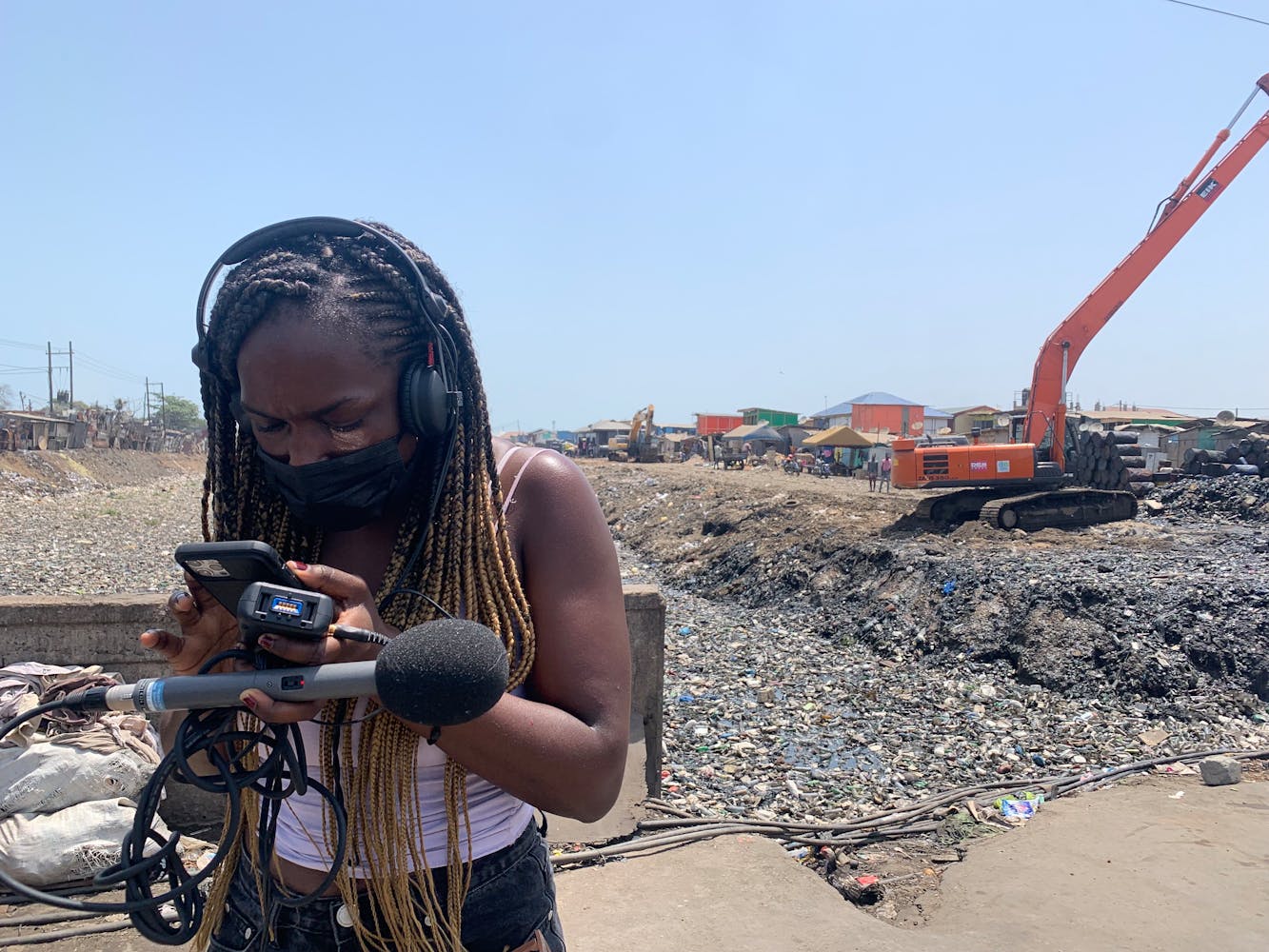 A women wearing a mask over her mouth uses a sound recorder and microphone while standing in an outdoor waste dump.