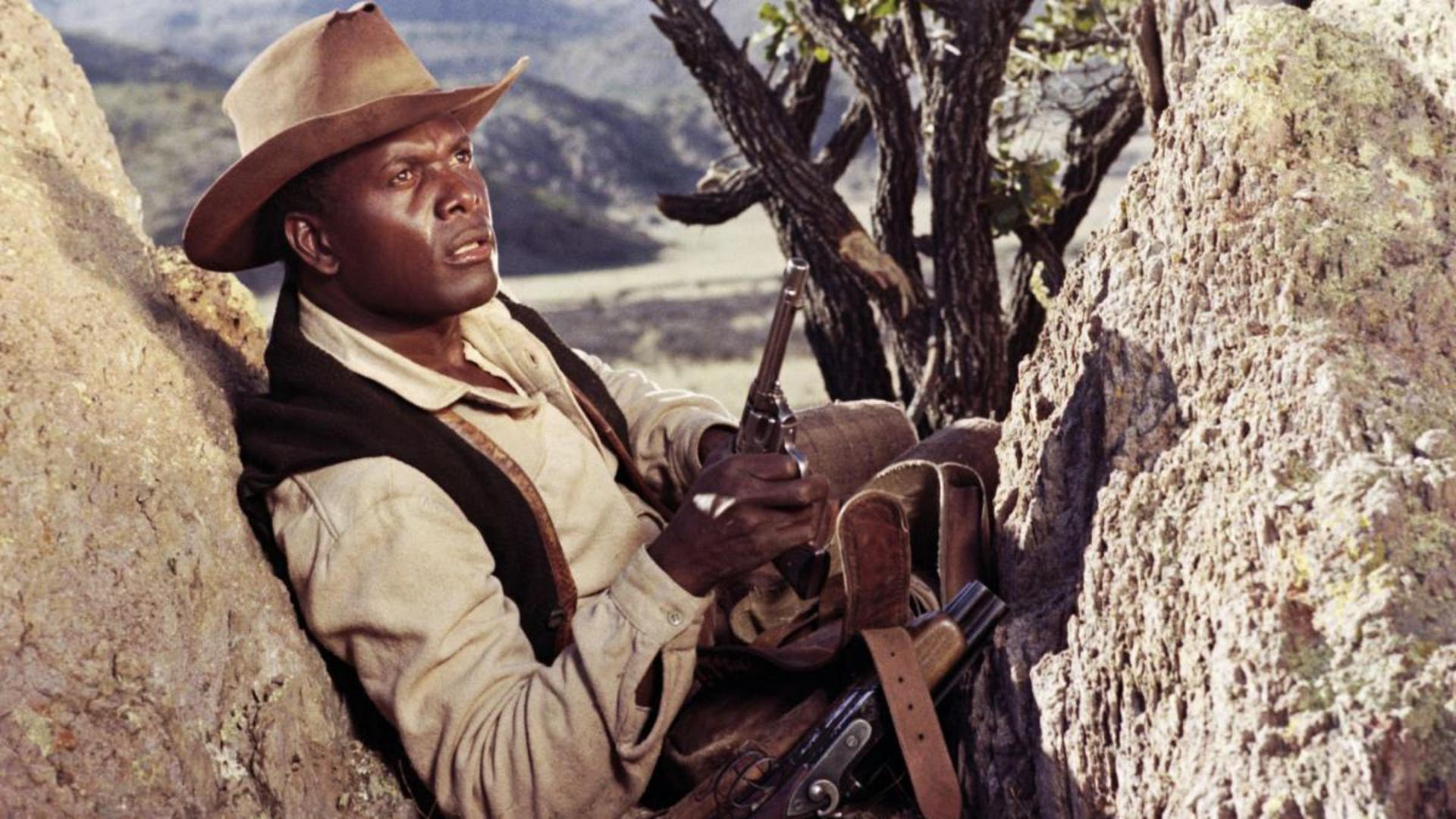 Sidney Poitier perches in between rocks and looks upward holding a handgun. He wears a cowboy hat and vest and has bags on his lap.