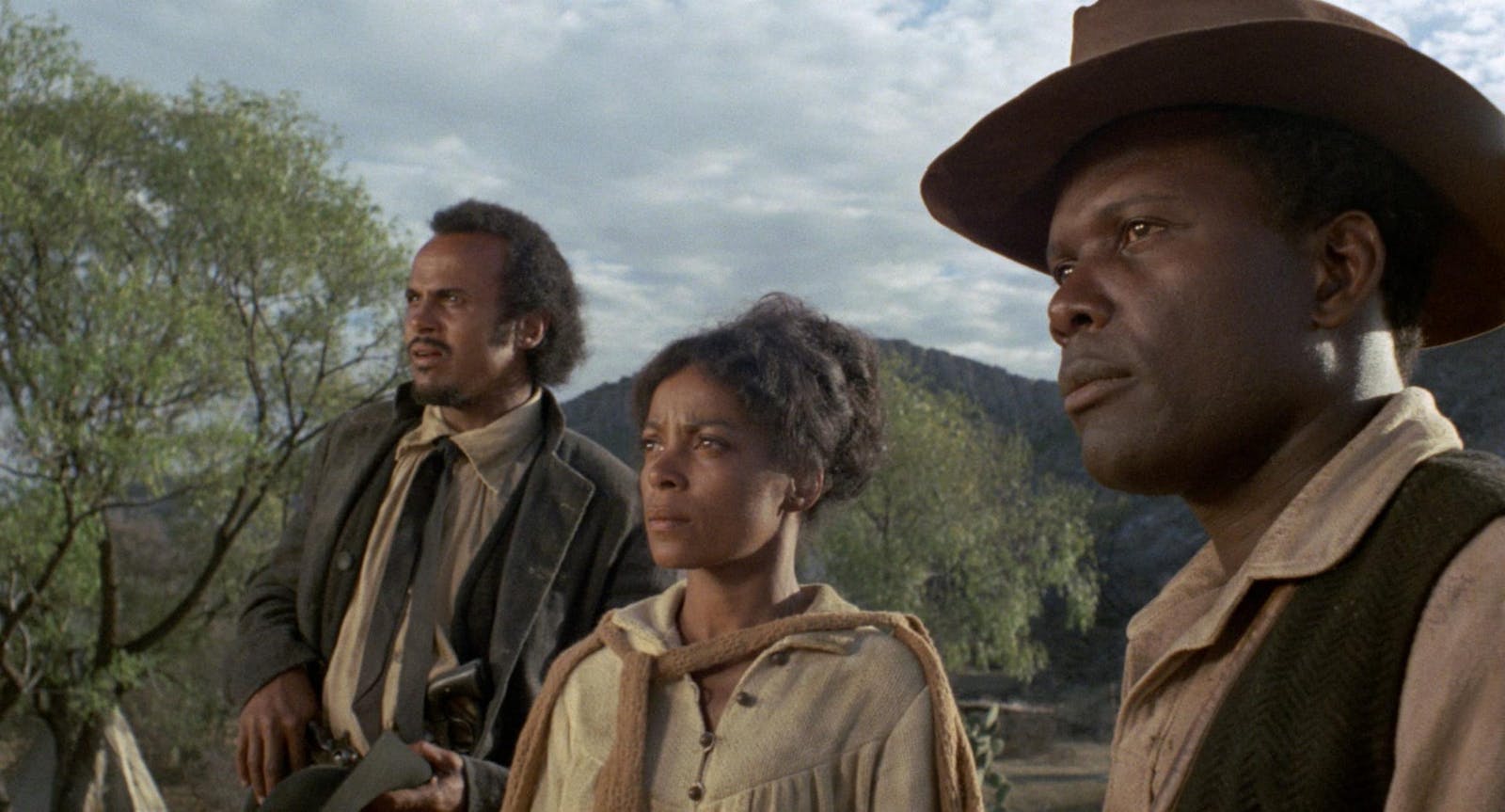 Three people with medium-dark skin tone look intently at something in the distance. They wear western clothing and there are mountains in the background.