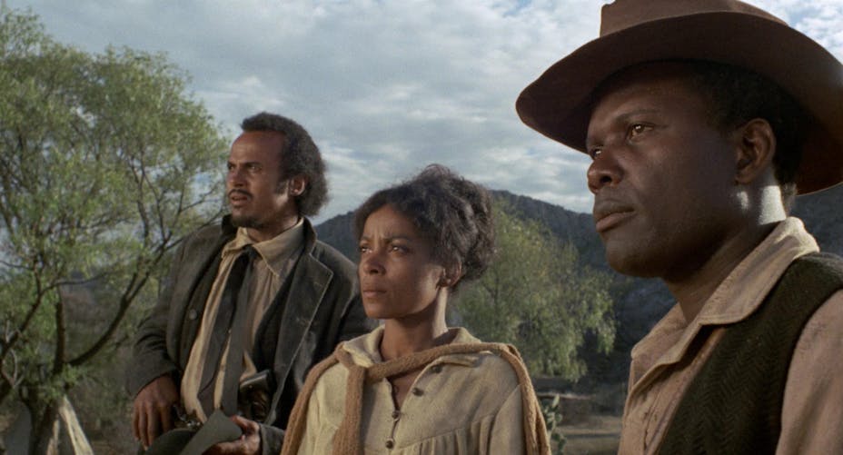 Three people with medium-dark skin tone look intently at something in the distance. They wear western clothing and there are mountains in the background.