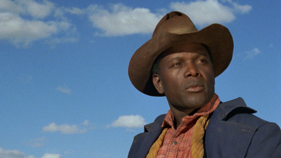 A close up of Sidney Poitier looking out toward the right, wearing a cowboy hat, a plaid shirt and blue jacket against a bright blue sky.