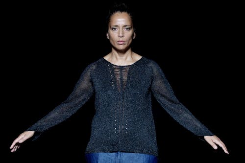 A person with medium-dark skin tone wears a slightly shimmery sweater, stares expressionless facing the camera, with their arms at a diagonal held at their sides.