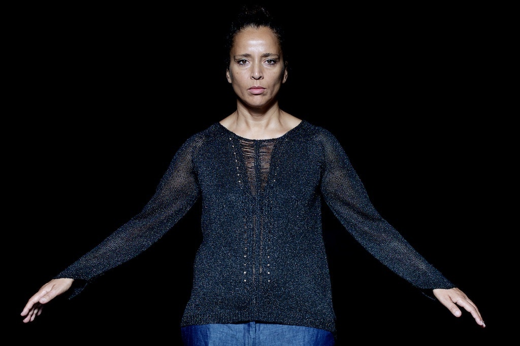 A person with medium-dark skin tone wears a slightly shimmery sweater, stares expressionless facing the camera, with their arms at a diagonal held at their sides.