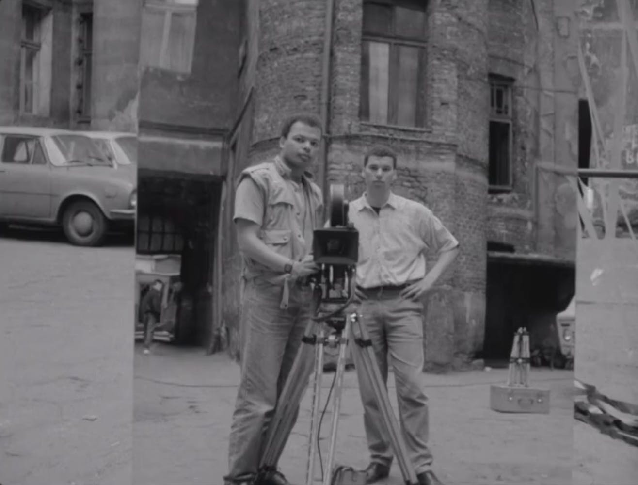 Two men standing next to a film camera on a city street looking at are reflected